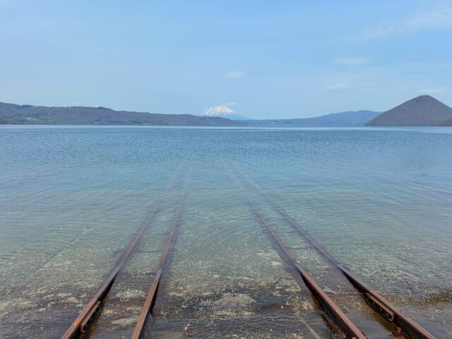A boat slipway running into the clear shallows of Lake Toya, with the far shore and a snow-capped cone on the horizon
