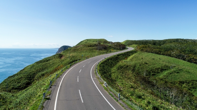A two-lane road curving along the top of green treeless sea cliffs, open water on the left and bare headlands running on ahead