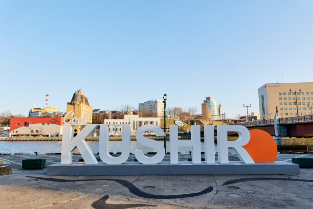 A large white and orange KUSHIRO sign standing on a riverside promenade, with the town, a red bridge and the water behind it